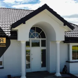 Elewacja domu jednorodzinnego z białymi oknami, drzwiami wejściowymi z półokrągłym przeszkleniem i czarną dachówką; widoczny pies siedzący przed oknem oraz logo firmy OKMAR Stolarka Budowlana...