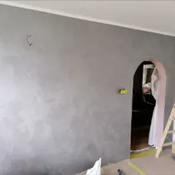 Ściana pokryta dekoracyjną szarą farbą z efektem strukturalnym, widoczna drabina malarska i zabezpieczona folią podłoga w trakcie prac wykończeniowych, zarysowane miejsce po gwoździu na ścianie.