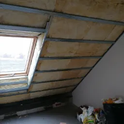 Skośny dach na poddaszu z oknem połaciowym, ocieplony wełną mineralną i przygotowany pod montaż płyt kartonowo-gipsowych, widoczny metalowy stelaż, na podłodze resztki materiałów budowlanych.
