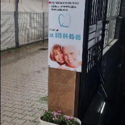 Tablica reklamowa laboratorium protetycznego z ofertą usług, numerem telefonu i uśmiechniętą parą, umieszczona przy wejściu do budynku, obok donicy z różowymi kwiatami.