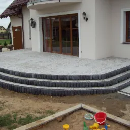 Półokrągły taras z jasnoszarej kostki brukowej z trzema stopniami obłożonymi ciemnoszarą kostką, przylegający do jasnego budynku z brązowymi drzwiami tarasowymi i oknem, w tle domy jednorodzinne,...