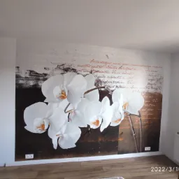 Tapeta z motywem białych orchidei i tekstu w odcieniach brązu, widoczna na całej ścianie, z fragmentem grzejnika i podłogi w dolnym rogu.