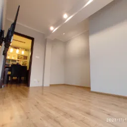 Salon z panelami podłogowymi w odcieniach drewna, ściany pomalowane na jasny szary kolor, na ścianie zamontowany telewizor.