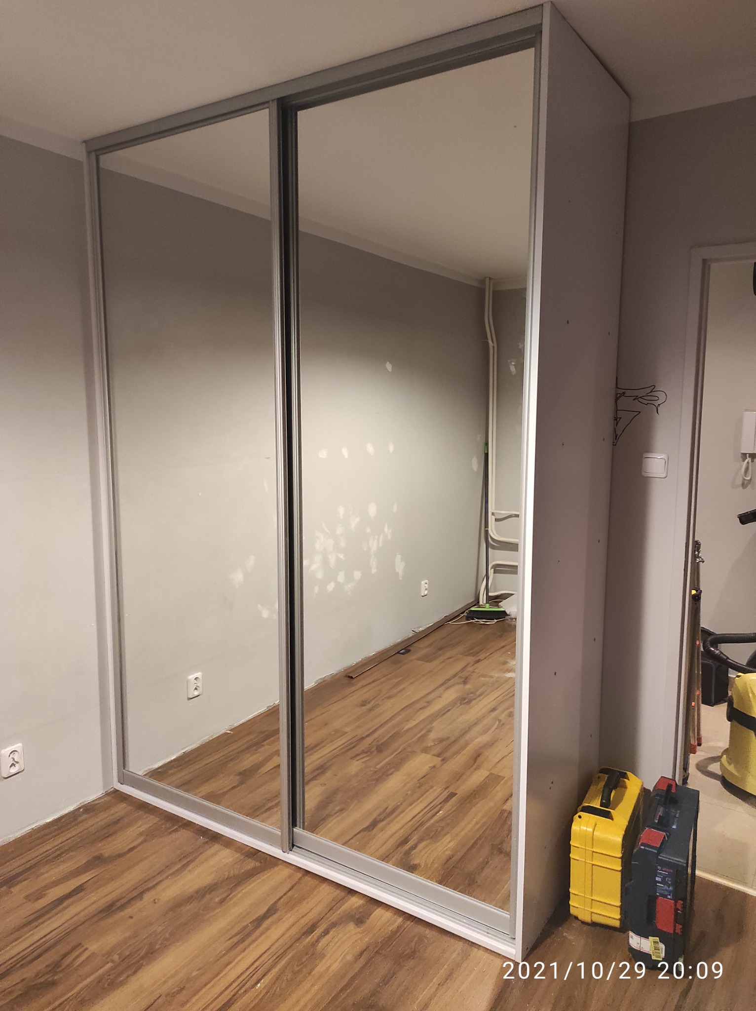 Wnętrze pokoju z zamontowaną szafą przesuwną z lustrami; ściany szare, częściowo z widocznymi śladami po naprawach, podłoga z paneli imitujących drewno, widoczne narzędzia w skrzynkach obok szafy.