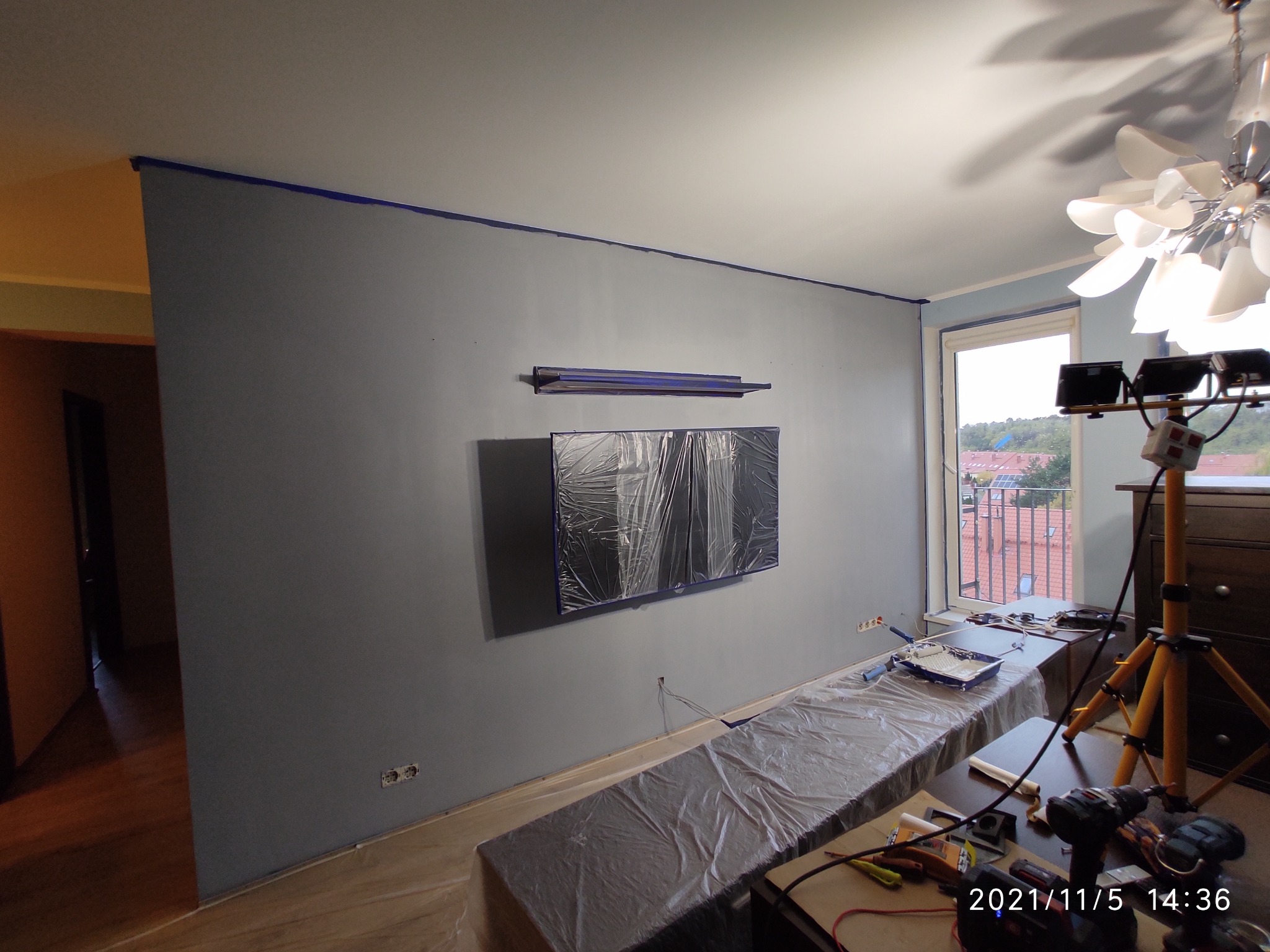 Przygotowanie do malowania pokoju: ściana zabezpieczona taśmą malarską, meble okryte folią ochronną, telewizor zabezpieczony folią, widoczny statyw oświetleniowy i narzędzia malarskie.