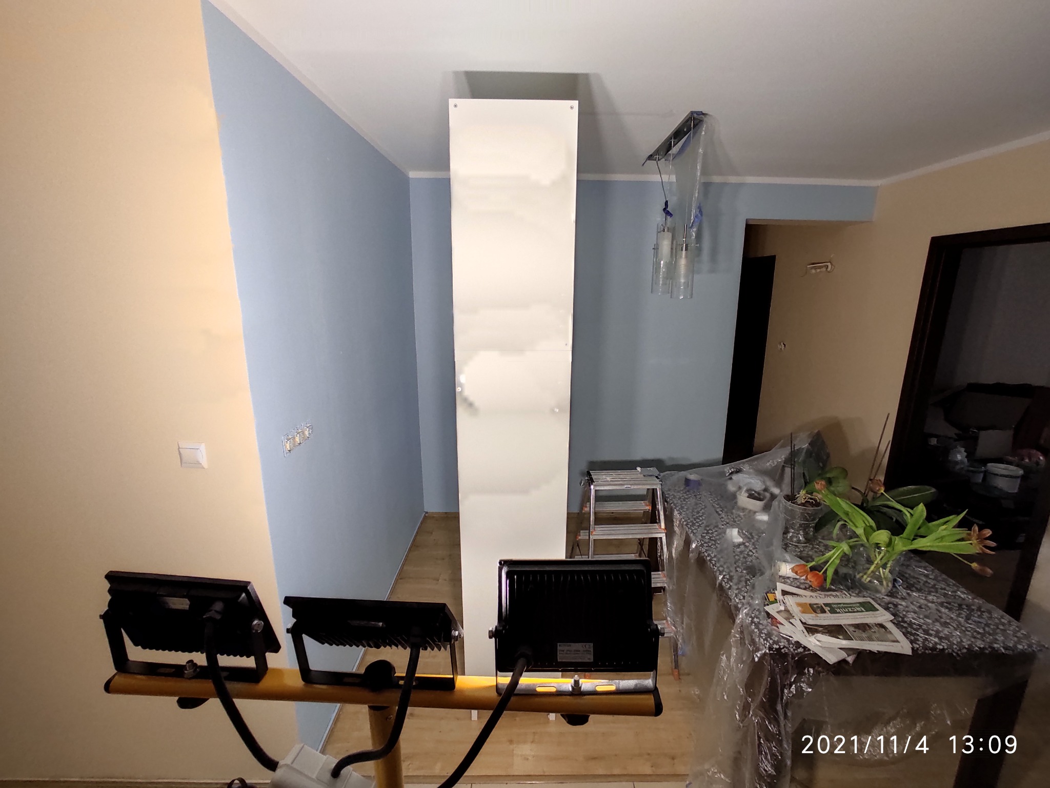 Wnętrze w trakcie prac malarskich: ściana pomalowana częściowo na niebiesko, biała kolumna, lampy halogenowe oświetlające przestrzeń, zabezpieczone meble folią ochronną.