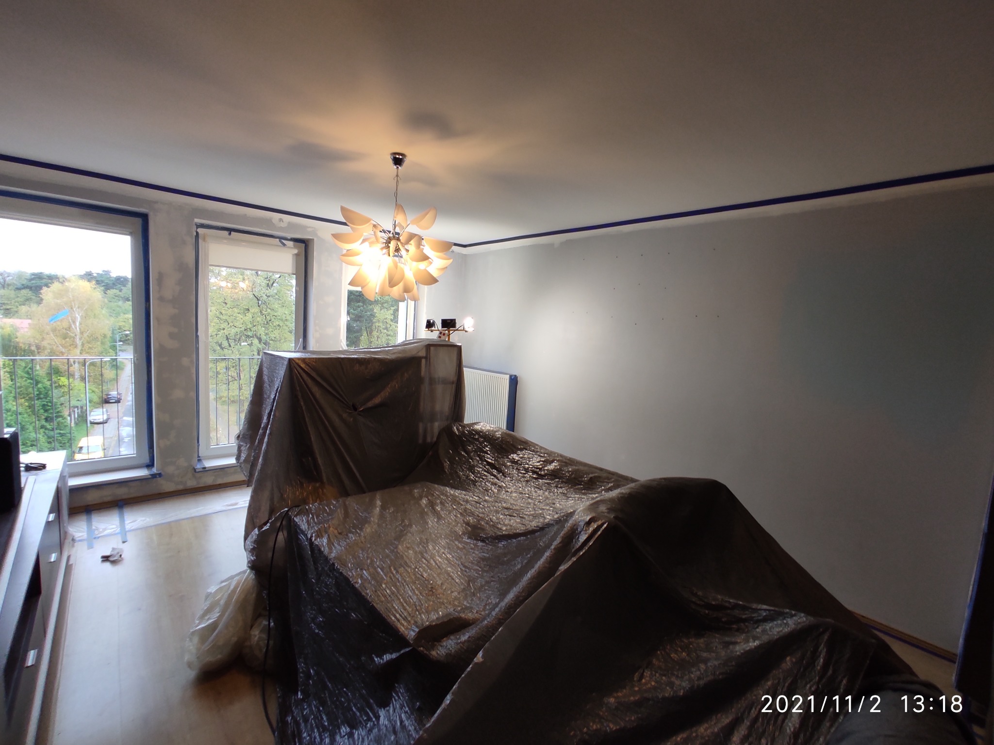 Pokój przygotowany do malowania: meble przykryte folią ochronną, ściany z widocznymi śladami szpachlowania i taśmą malarską przy suficie i oknach.