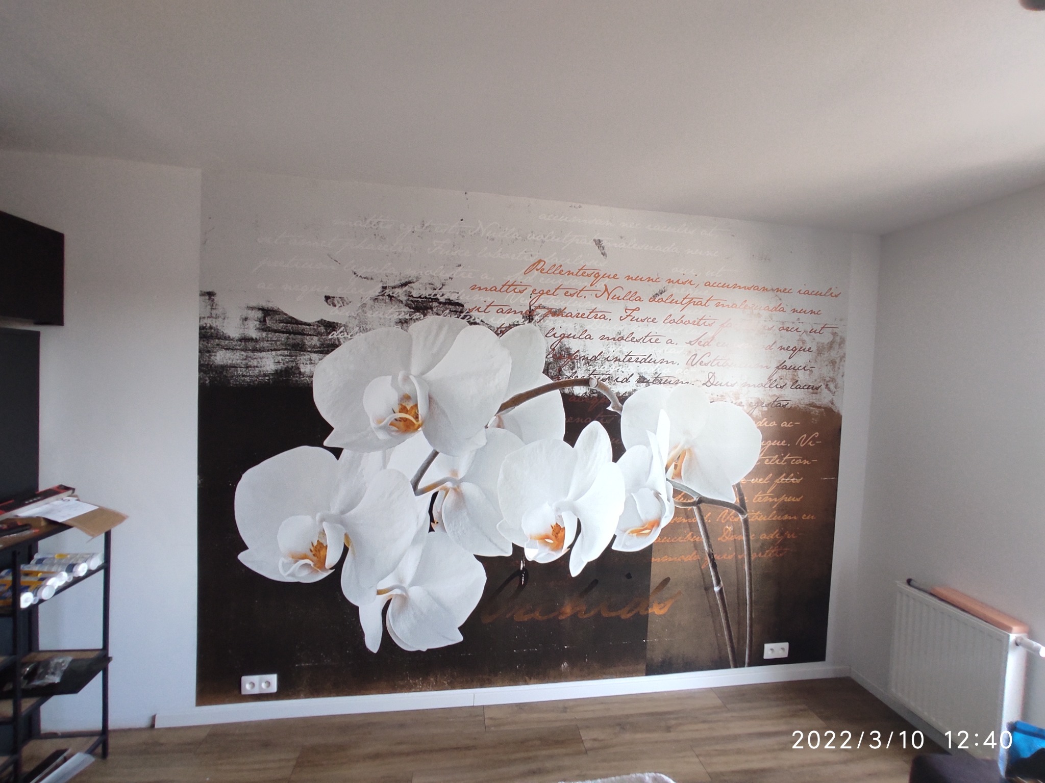 Tapeta z motywem białych orchidei i tekstu w odcieniach brązu, widoczna na całej ścianie, z fragmentem grzejnika i podłogi w dolnym rogu.