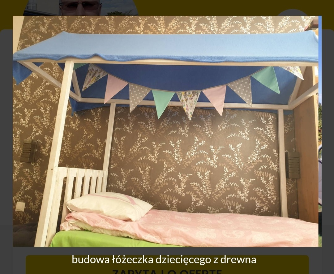 Drewniane łóżeczko dziecięce z baldachimem z niebieskiego materiału i girlandą z trójkątnych chorągiewek na tle tapety w roślinne wzory.