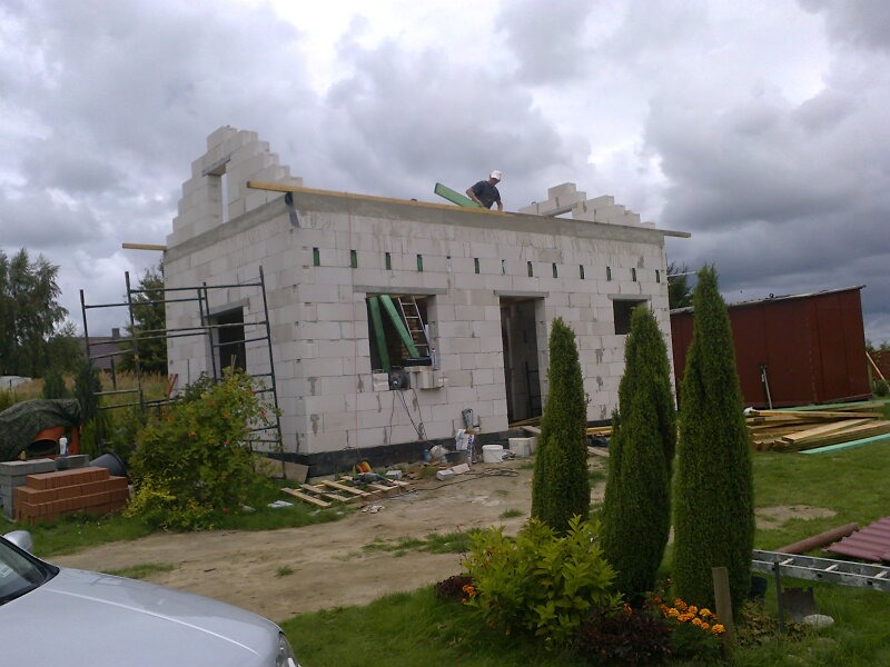 Budowa domu jednorodzinnego w stanie surowym otwartym, widoczne ściany z bloczków, częściowo wykonana więźba dachowa oraz pracownik na dachu.