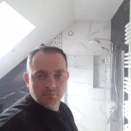 Ukończona łazienka na poddaszu ze skośnym oknem dachowym, kabina prysznicowa z marmurowymi płytkami, widoczny mężczyzna w czarnej bluzie robiący selfie.