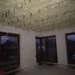 Poddasze w trakcie ocieplania pianą poliuretanową, widoczne metalowe elementy mocujące, okna z ciemnymi szybami, surowe ściany i podłoga.