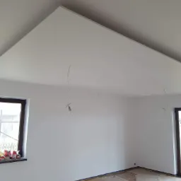 Wnętrze pokoju po świeżym pomalowaniu na biało, widoczne zabezpieczone okno z farbami, wystający kabel z żar&oacute;wką i fragment podłogi zabezpieczony kartonem.