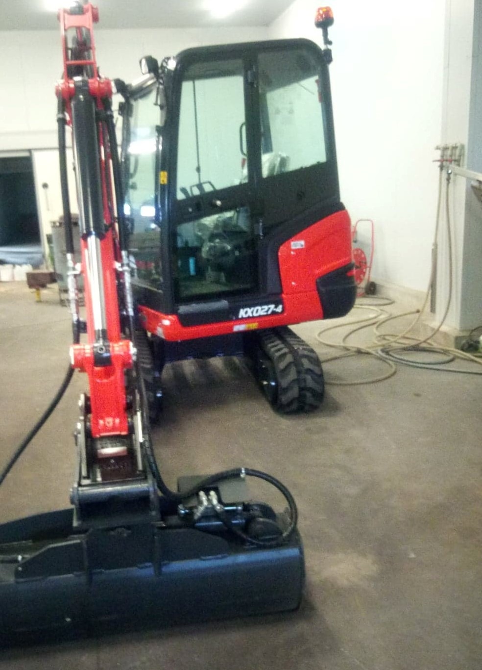 Czerwono-czarna mini koparka Kubota KX027-4 na gąsienicach, stojąca w jasnym pomieszczeniu z betonową podłogą i białymi ścianami, widoczne przewody i narzędzia w tle.