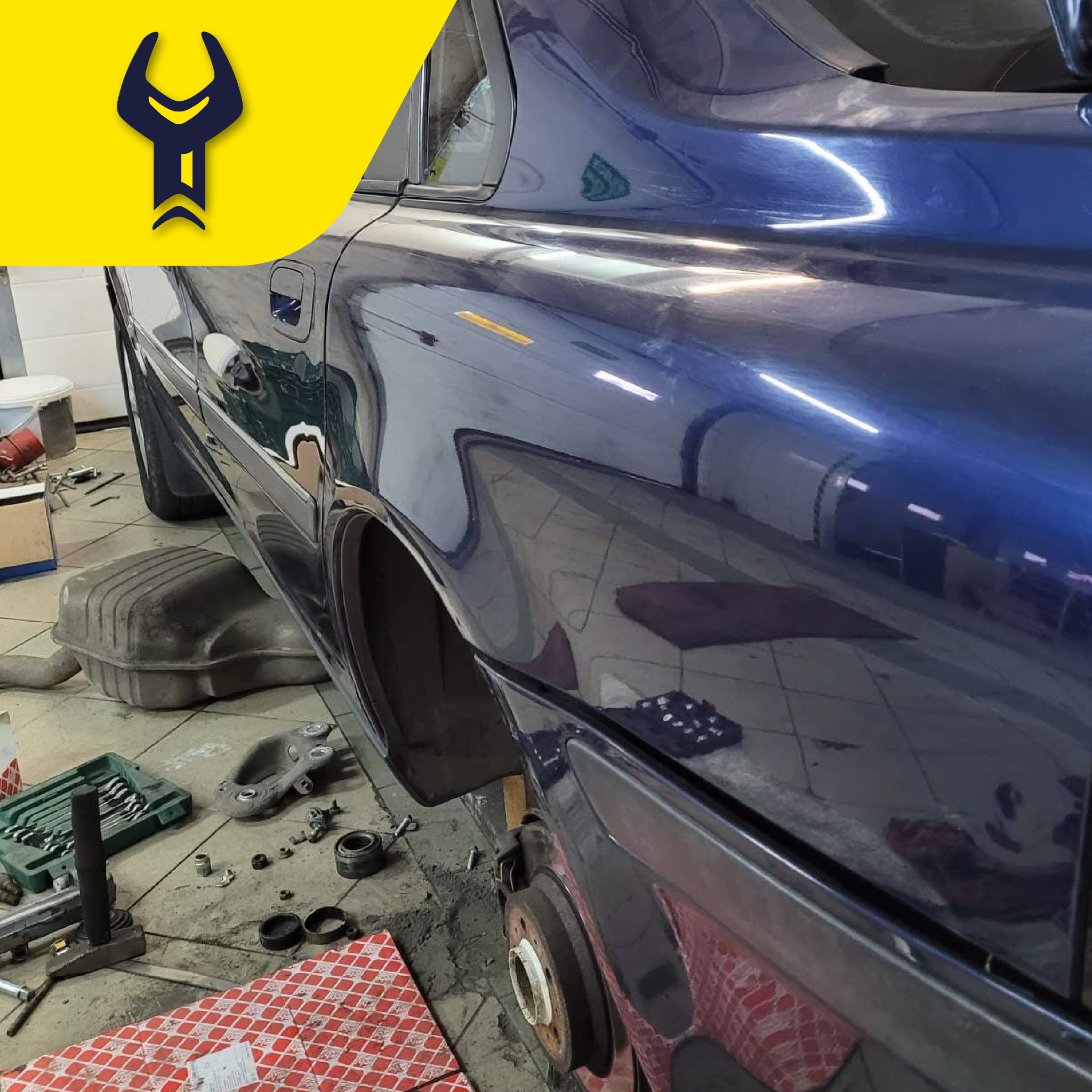 Niebieski samochód bez koła, w warsztacie, widoczne narzędzia, klucze, podnośnik, element zawieszenia oraz tłumik na podłodze.