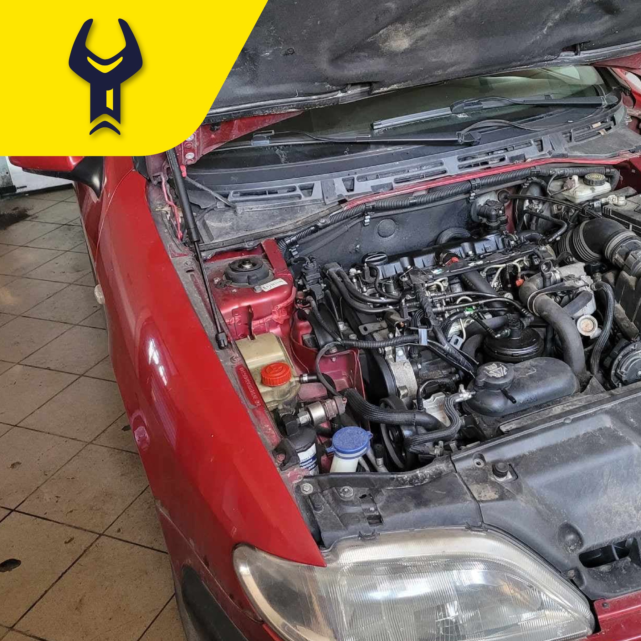 Otwarta maska czerwonego samochodu, widoczny silnik z licznymi przewodami i elementami, na pierwszym planie reflektor, tło stanowi podłoga warsztatu.