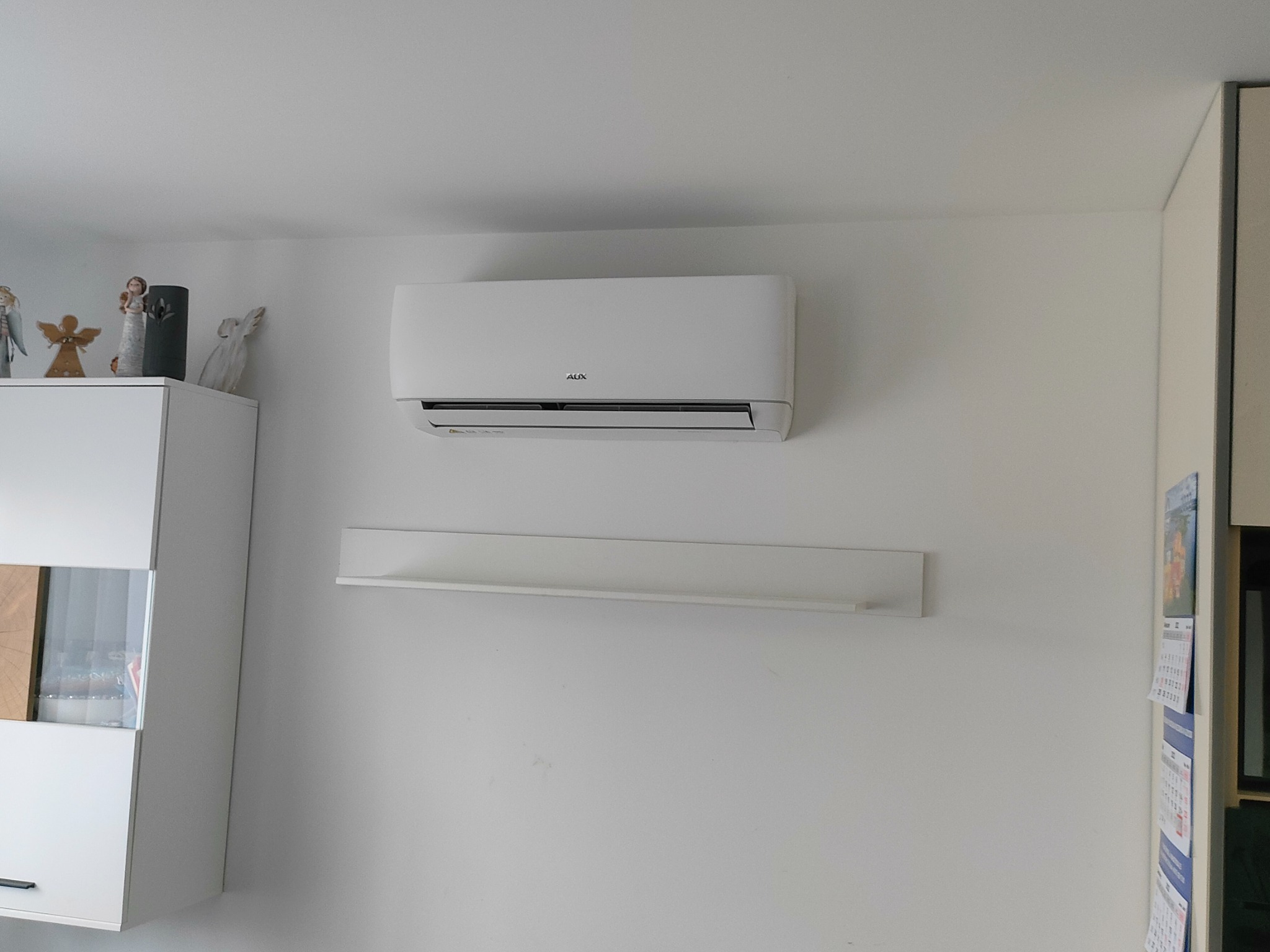 Biały klimatyzator marki AUX zamontowany na ścianie w jasnym pomieszczeniu, pod nim wąska biała półka. Obok biały mebel z figurkami aniołów na górze.