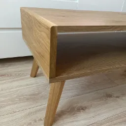 Stolik kawowy z jasnego drewna z półką, na skośnych nogach, ustawiony na panelach podłogowych imitujących drewno, z białymi frontami meblowymi w tle.