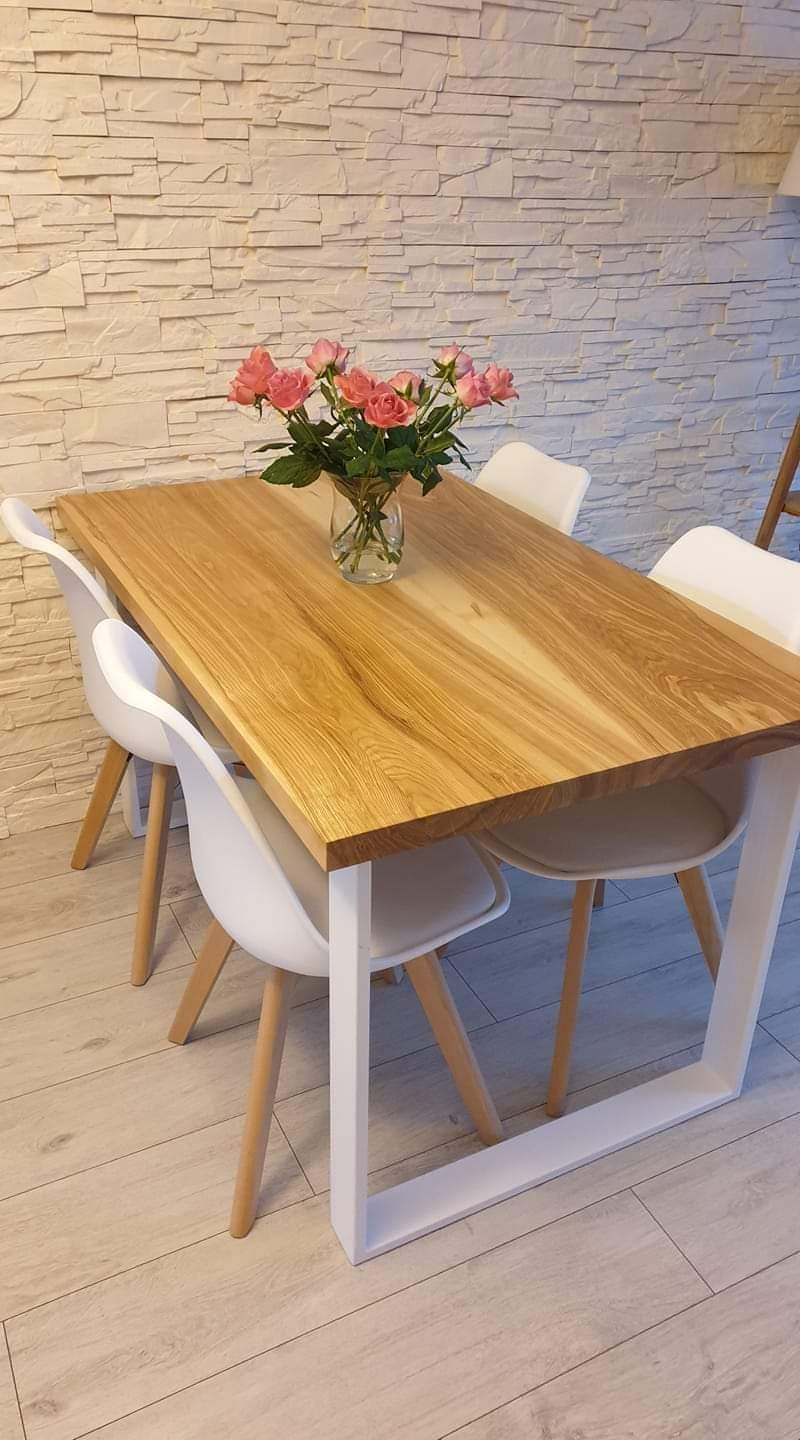 Stół jadalny z blatem z jasnego drewna i białymi nogami w kształcie prostokąta, otoczony białymi krzesłami, na blacie wazon z różowymi różami, w tle ściana z dekoracyjnej cegły.