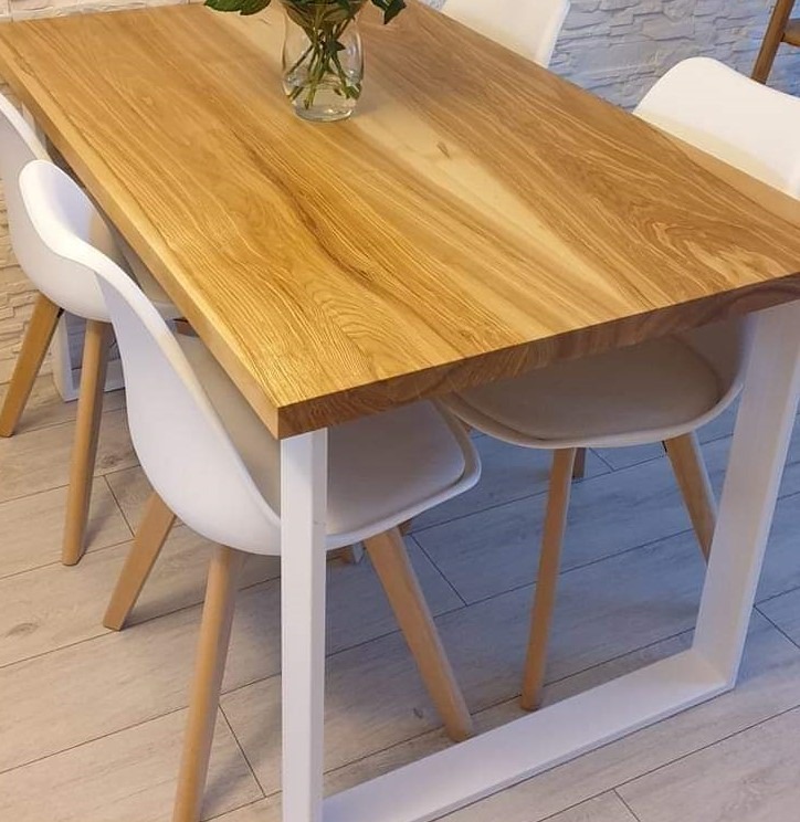 Stół z blatem z jasnego drewna o wyraźnym usłojeniu, wsparty na białych, metalowych nogach w kształcie litery U, otoczony białymi krzesłami z drewnianymi nogami, na podłodze z paneli.