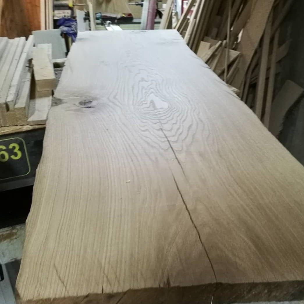 Surowy, jasny blat z litego drewna z wyraźnym usłojeniem i naturalnymi pęknięciami, leżący w warsztacie.