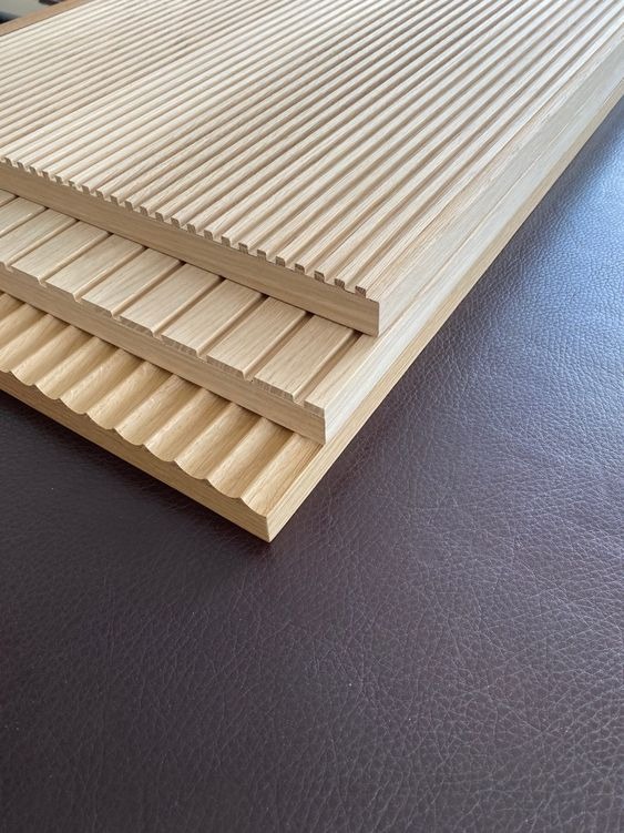 Trzy ułożone deski z jasnego drewna z frezowanymi wzorami: regularne rowki, szerokie żłobienia i falista krawędź, leżące na ciemnobrązowej, skórzanej powierzchni.