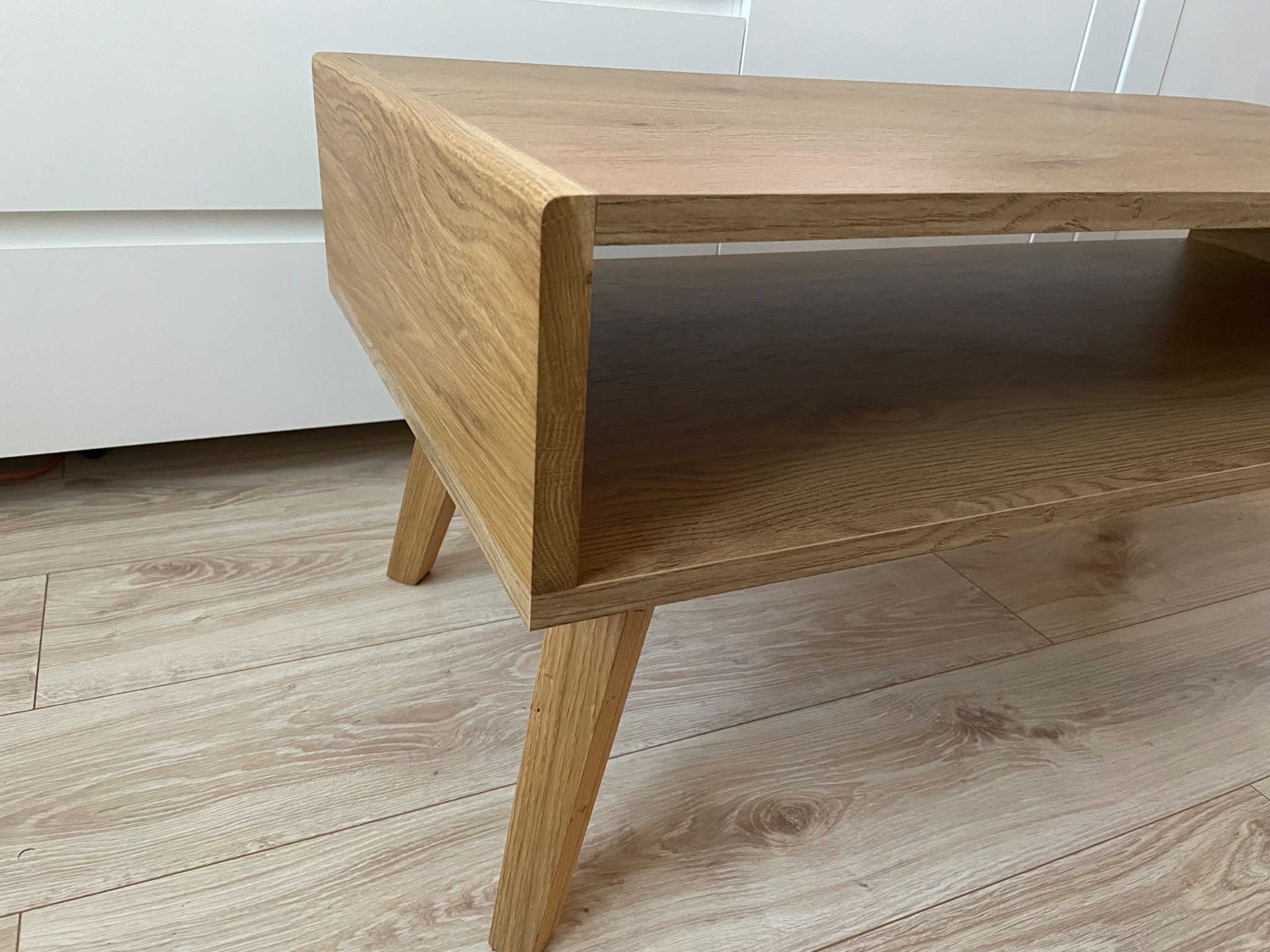 Stolik kawowy z jasnego drewna z półką, na skośnych nogach, ustawiony na panelach podłogowych imitujących drewno, z białymi frontami meblowymi w tle.