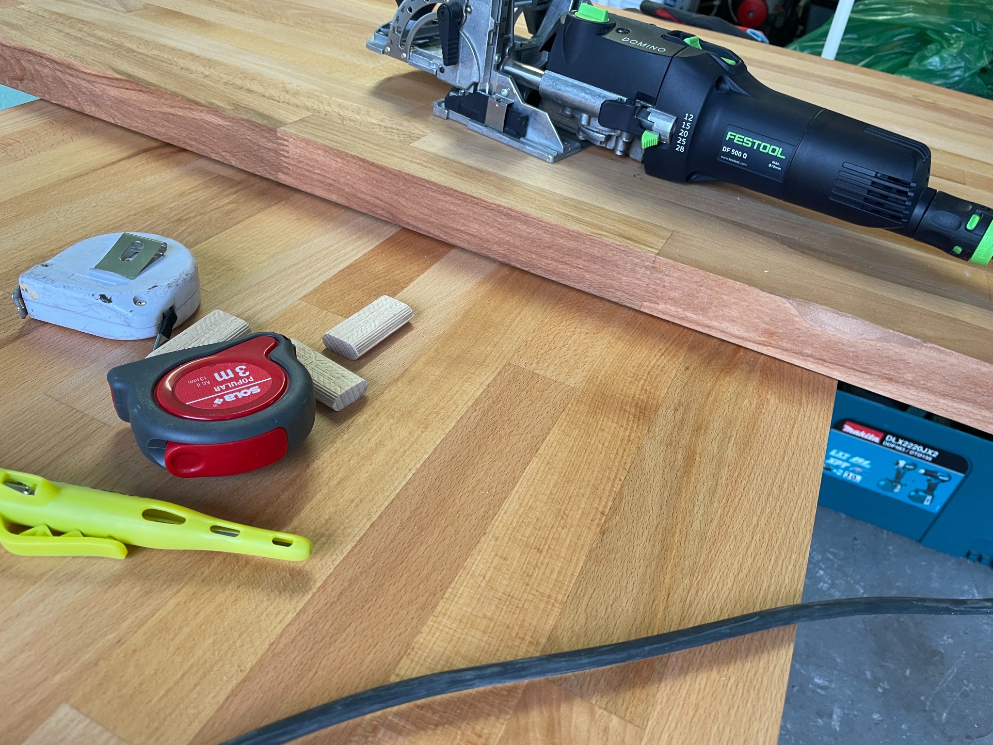 Przygotowanie do łączenia drewnianego elementu za pomocą frezarki Festool Domino, widoczne kołki, miara zwijana Sola, oraz żółty zacisk stolarski na blacie roboczym.