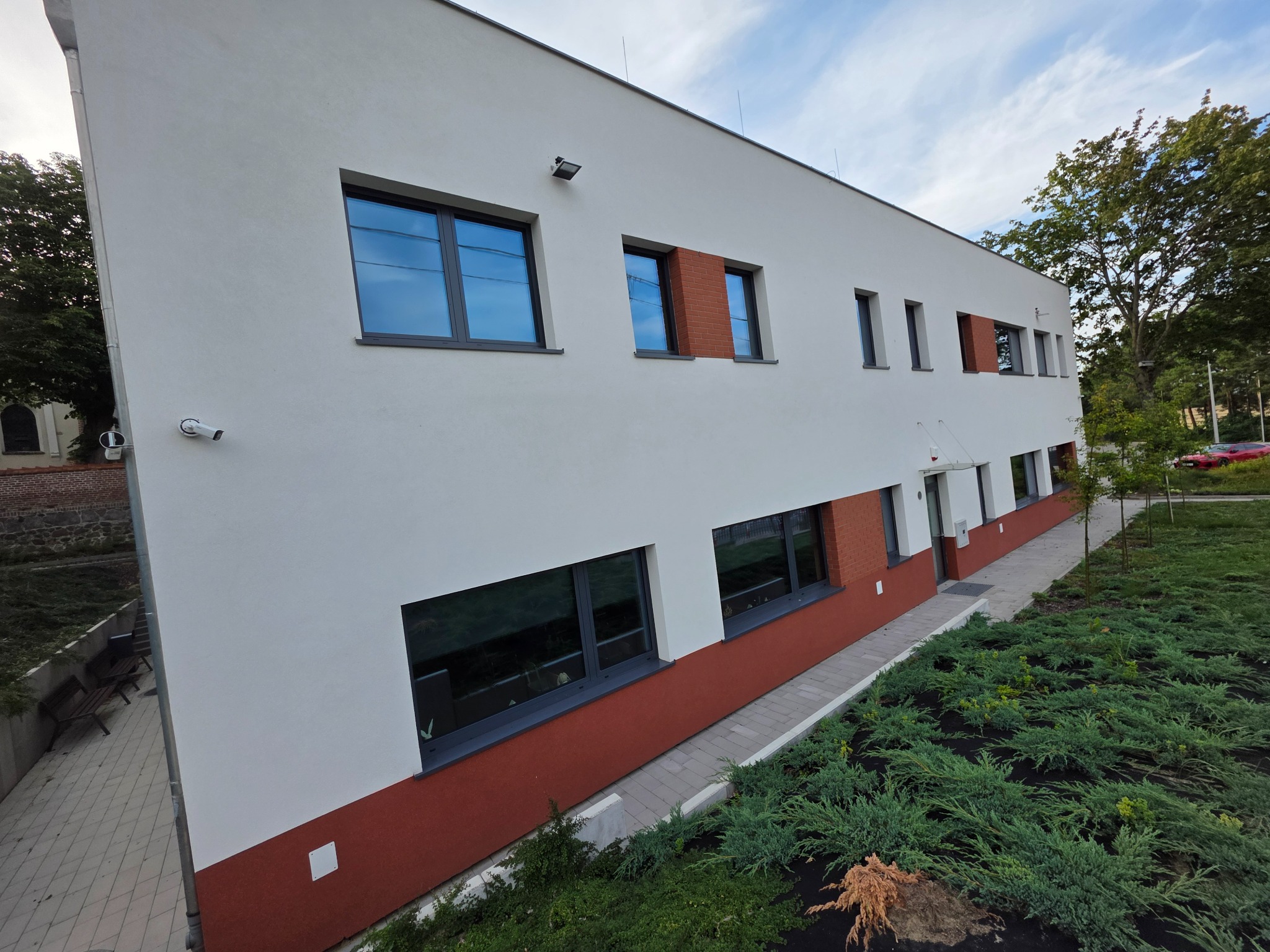 Nowoczesny budynek z białą elewacją, szarymi oknami i ceglanymi akcentami. Widoczne elementy architektury i otoczenie budynku w słoneczny dzień.