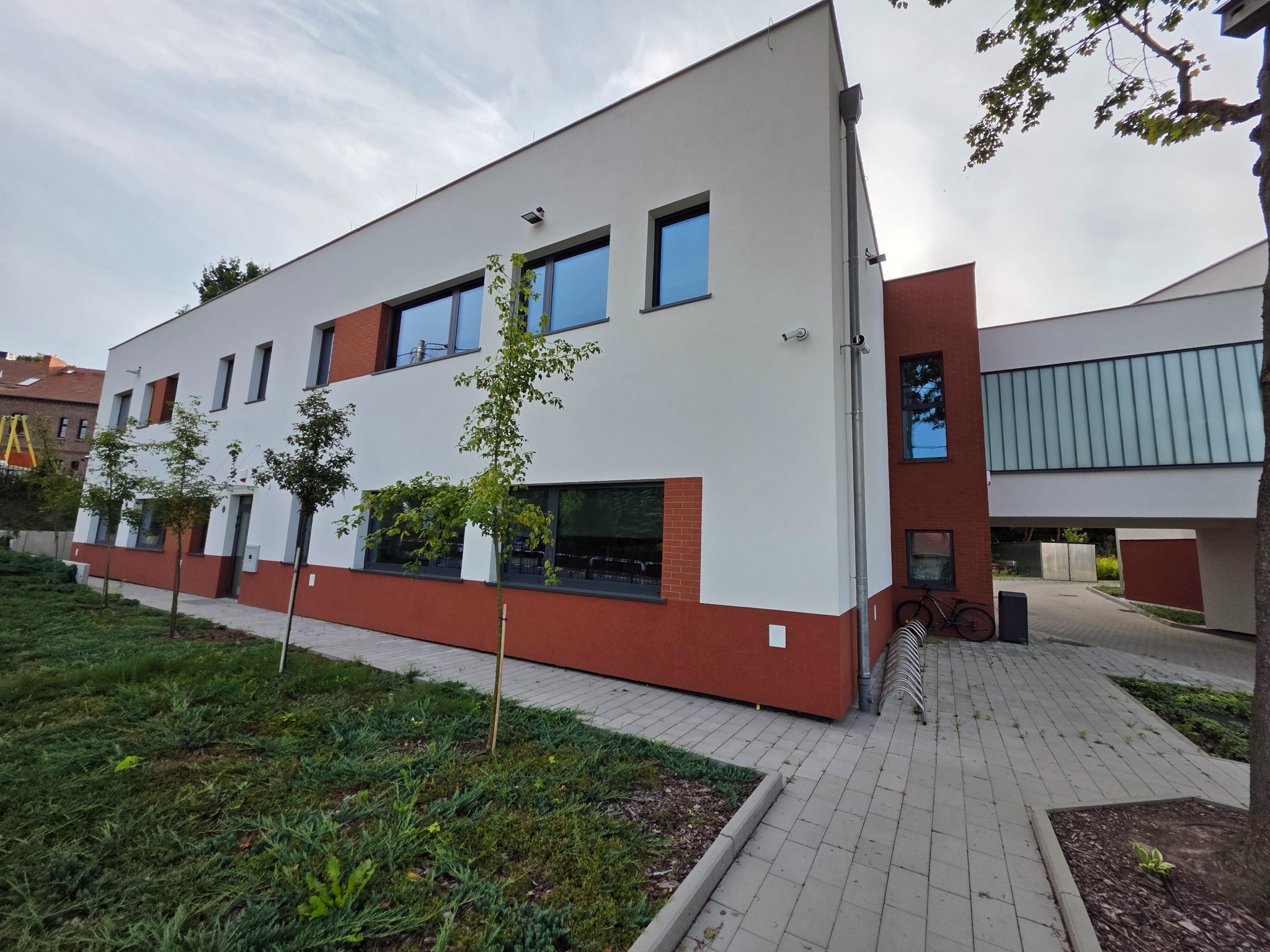 Dwukondygnacyjny budynek z białymi ścianami i czerwonymi akcentami, małe drzewka przed budynkiem, widok z dołu. Nowoczesna architektura.