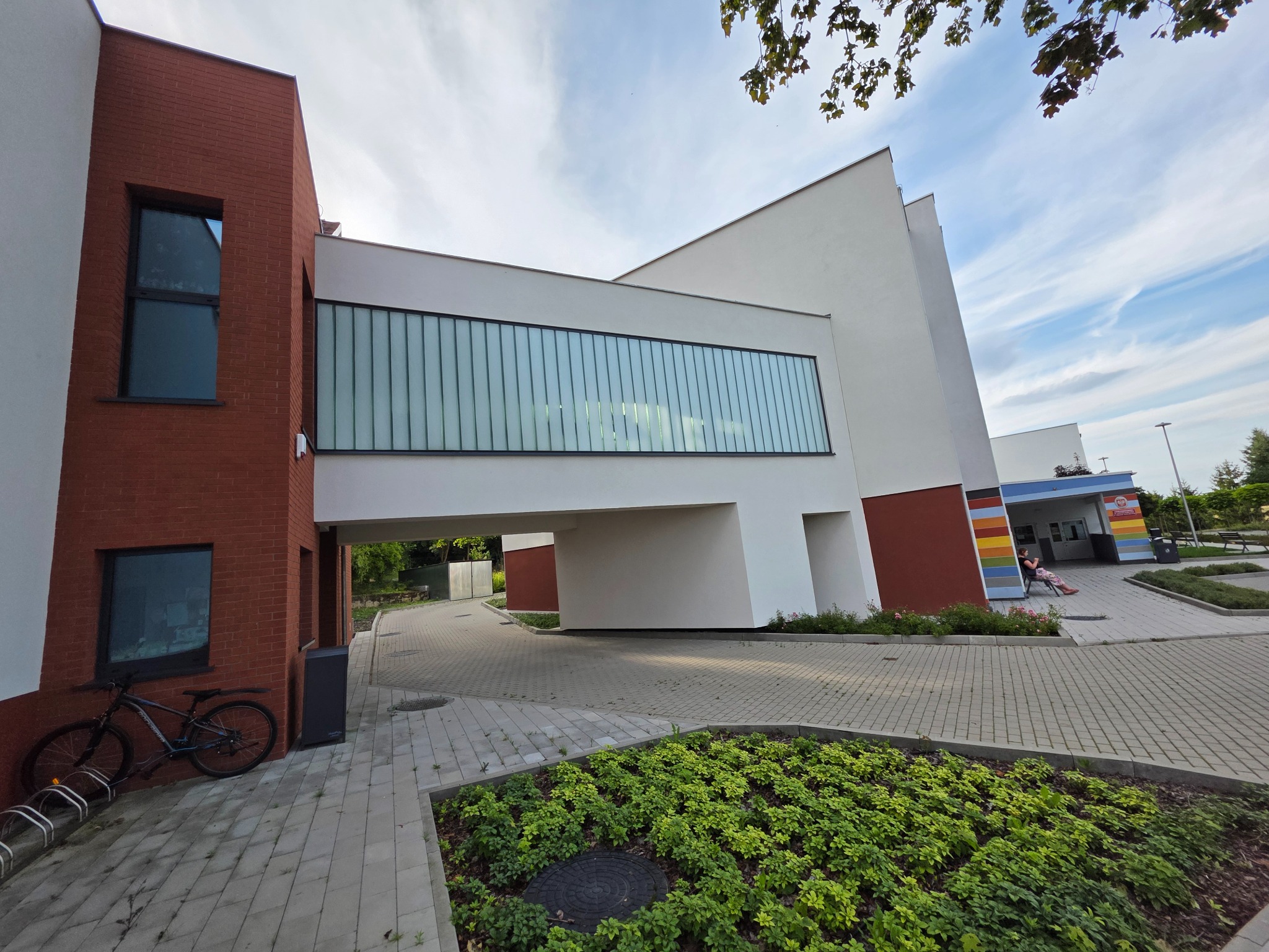Nowoczesny budynek z czerwonej cegły i białych paneli, połączony przejściem nad chodnikiem. W tle widoczne drzewa i fragment nieba.
