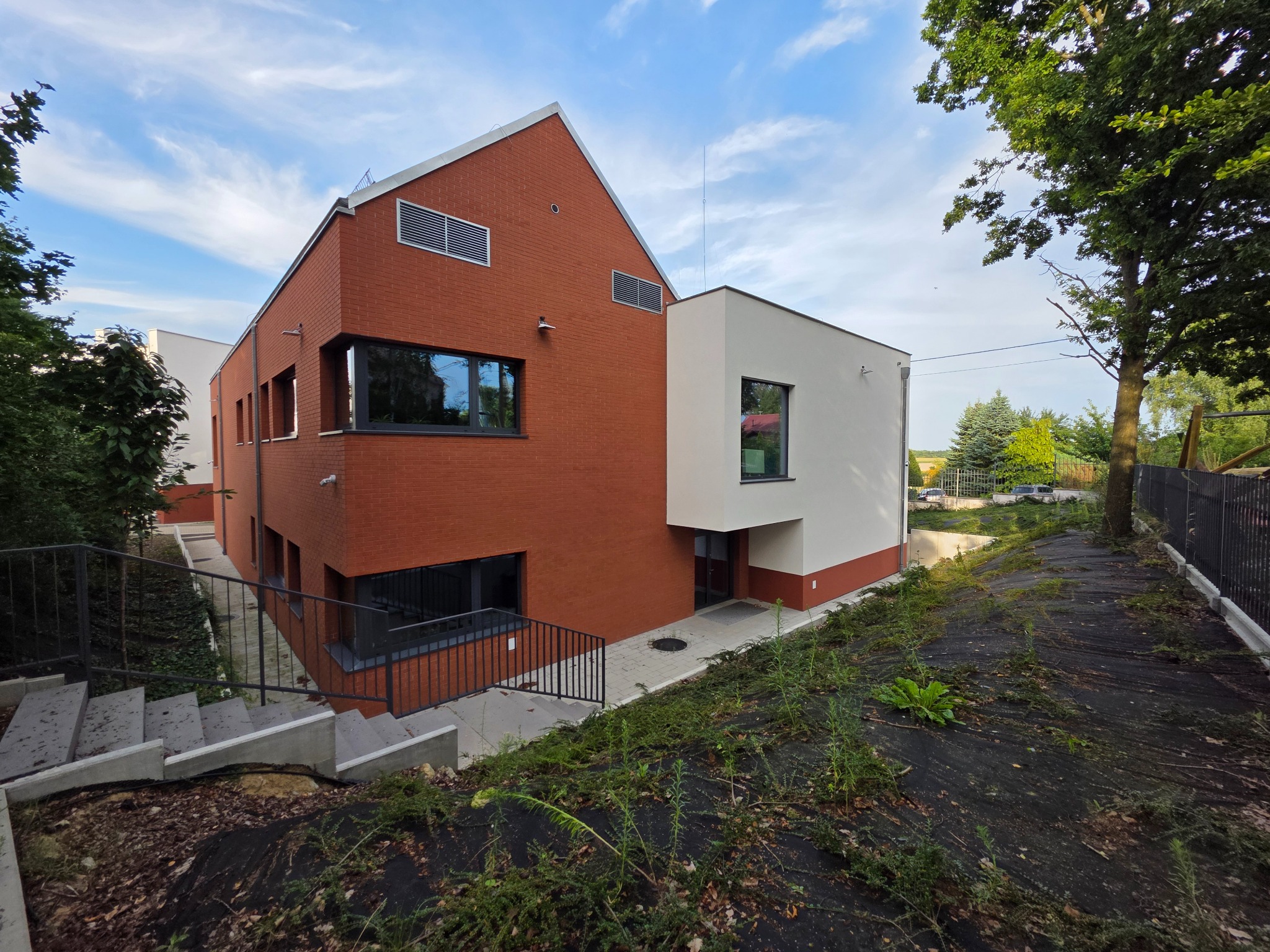 Nowoczesny budynek z ceglaną elewacją i białym aneksem, widoczny z dołu, z zielenią w tle i schodami prowadzącymi do wejścia.