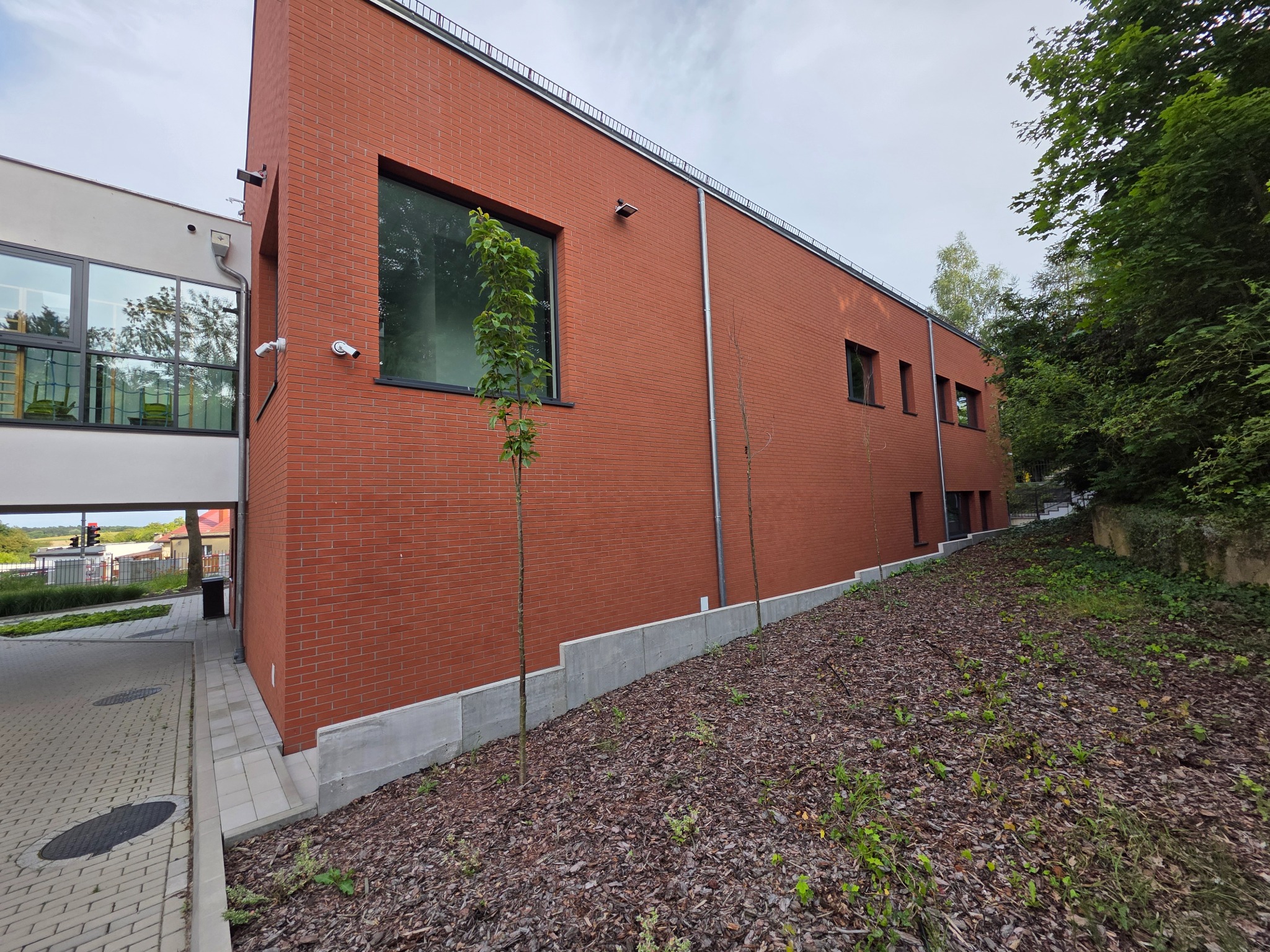 Ceglana elewacja budynku z nowoczesnymi oknami i systemem kamer. Widoczne fragmenty chodnika i zieleni.