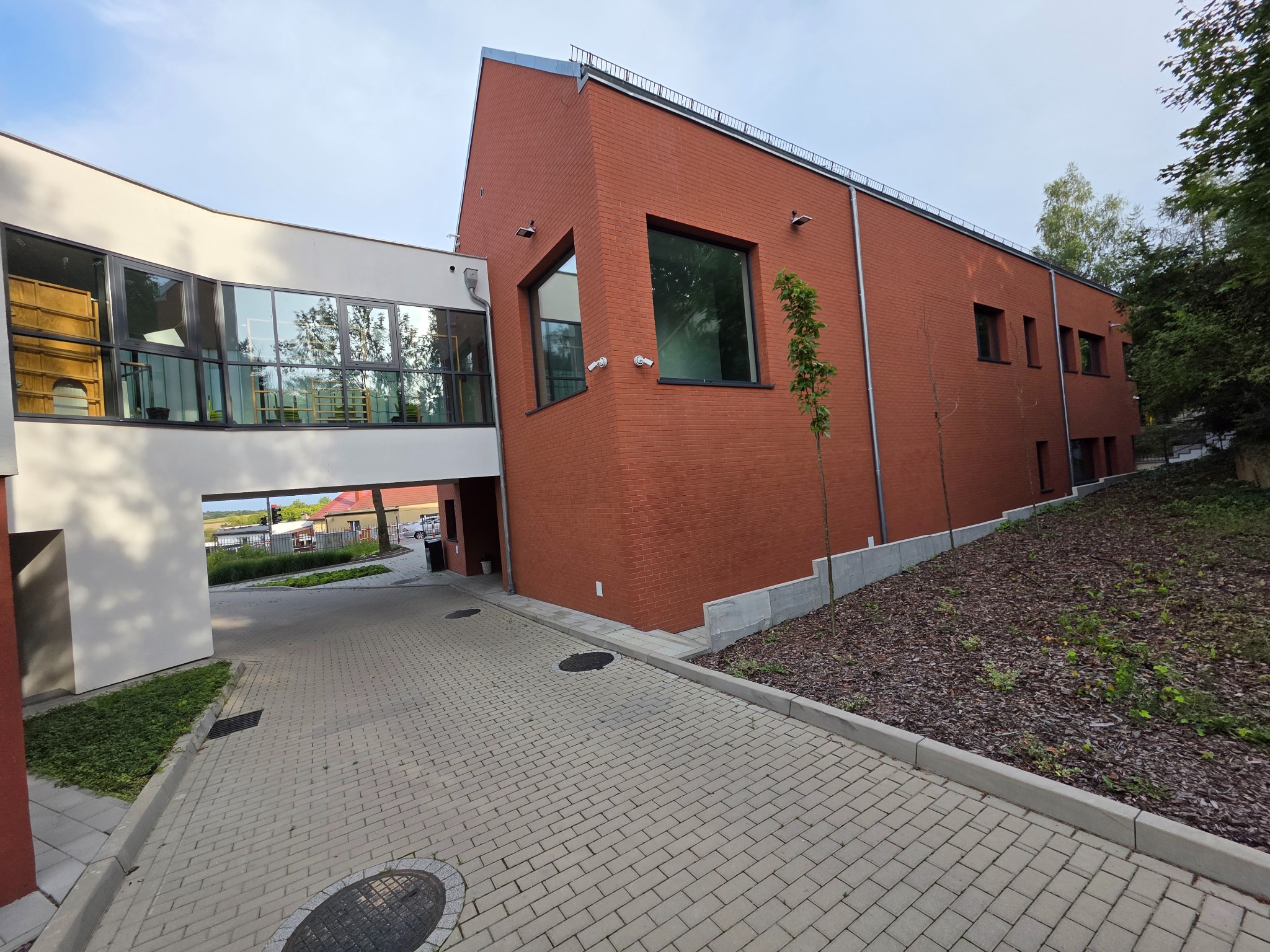 Nowoczesny budynek z czerwonej cegły, połączony przeszklonym łącznikiem, widoczny z brukowanego podjazdu. Uwagę zwracają geometryczne kształty i minimalistyczny design.