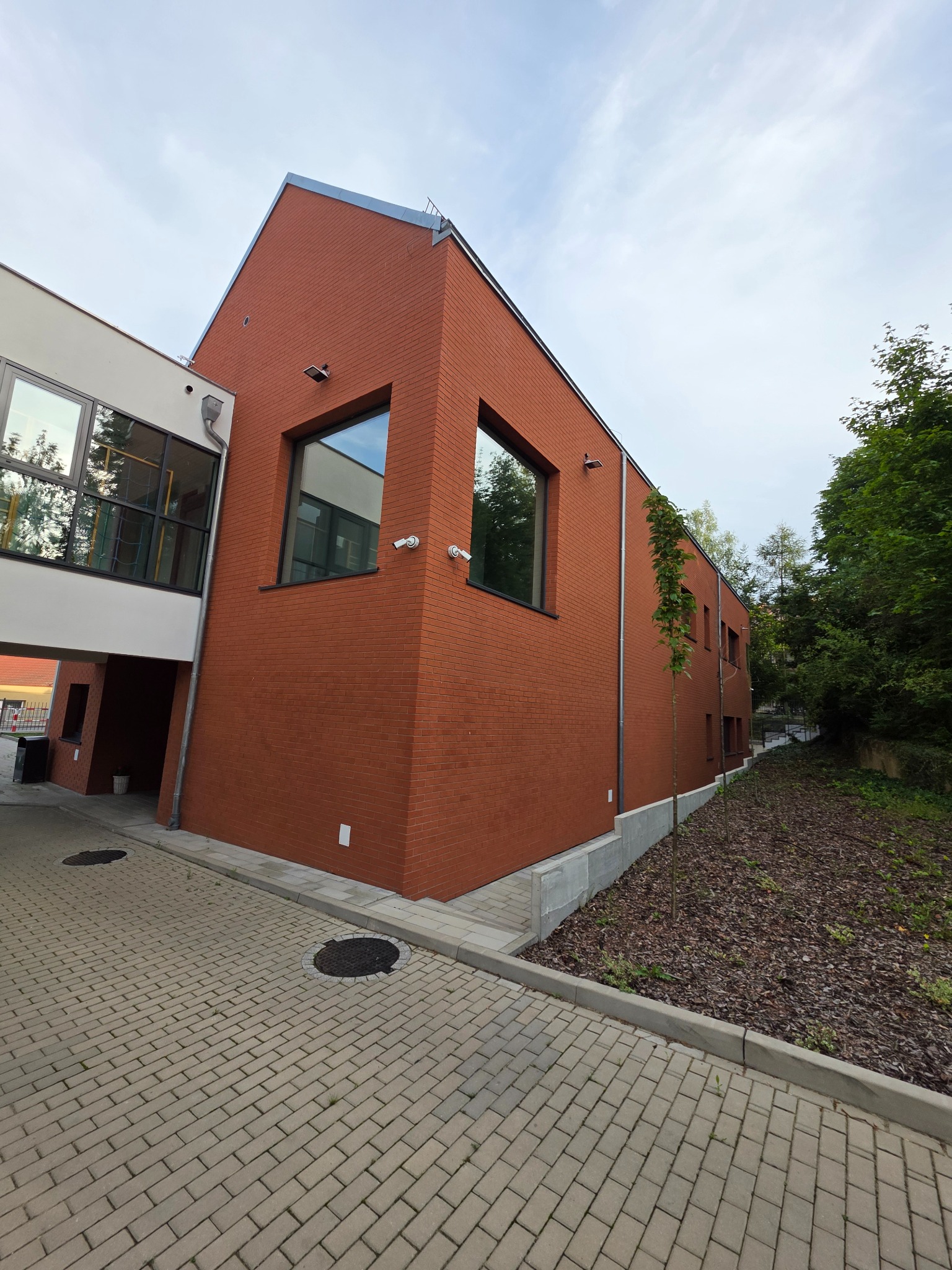 Nowoczesny budynek z cegły klinkierowej w odcieniach czerwieni, z widocznymi oknami i systemem kamer monitoringu, otoczony brukowanym chodnikiem i zielenią.