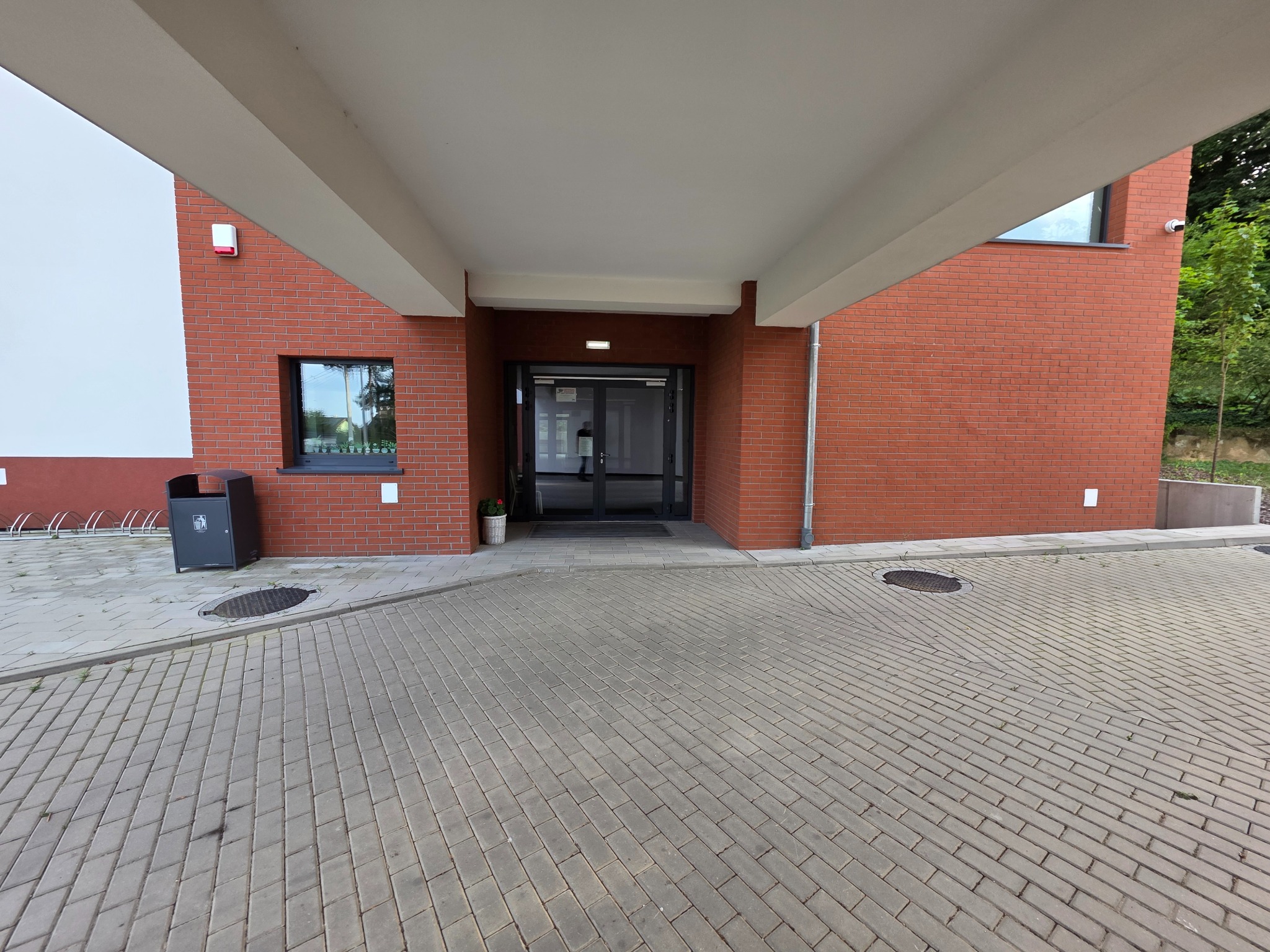 Nowoczesny budynek z ceglaną elewacją i szarym chodnikiem z kostki. Widoczne drzwi wejściowe, okno i elementy wykończeniowe. Minimalistyczny design.