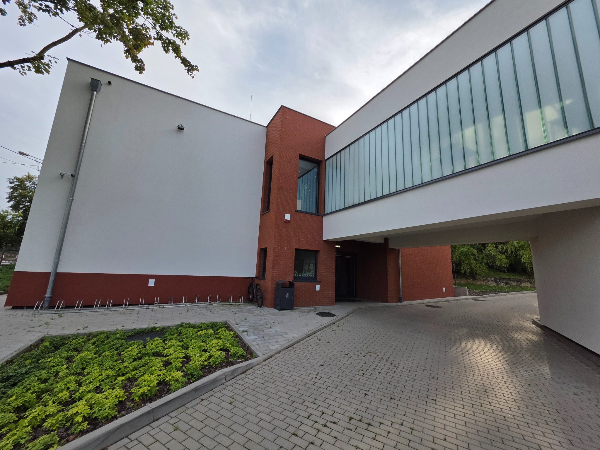 Nowoczesny budynek z białymi i ceglanymi elementami elewacji, połączony szklanym łącznikiem. Widoczny chodnik z kostki brukowej i zieleń.