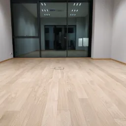 Jasne, drewniane panele ułożone w nowym biurze, widoczne duże okno z czarną ramą i minimalistyczne wykończenie.