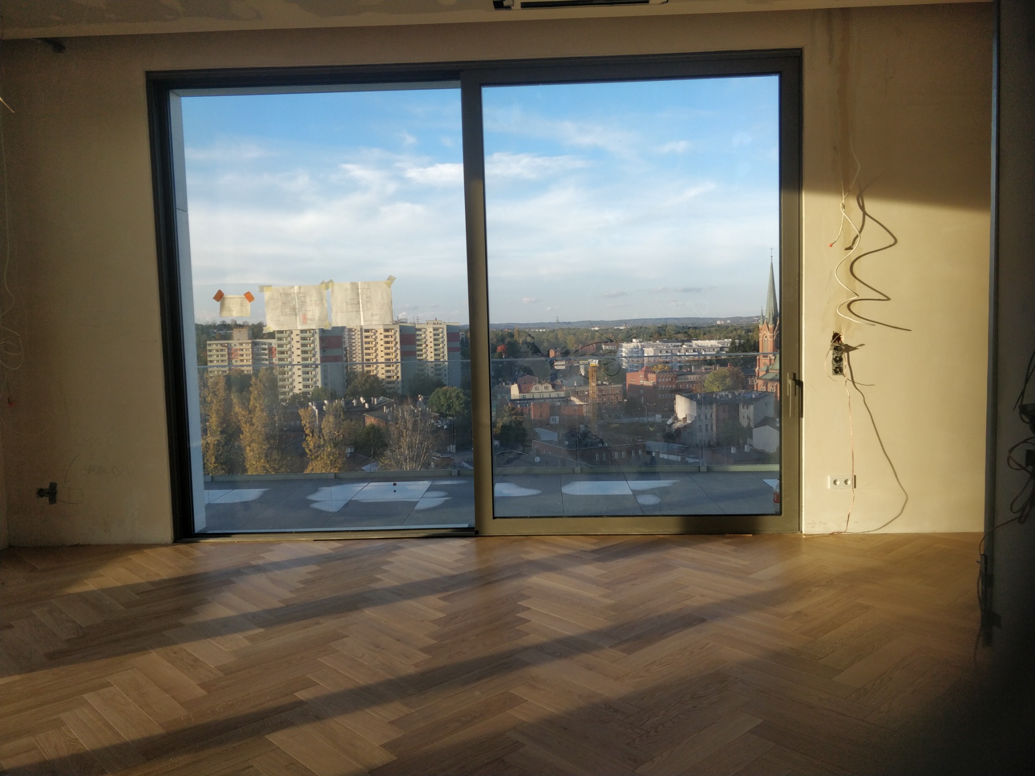 Parkiet ułożony w jodełkę francuską w jasnym pomieszczeniu z dużym oknem balkonowym i widokiem na miasto. Na ścianie widoczne przewody elektryczne.