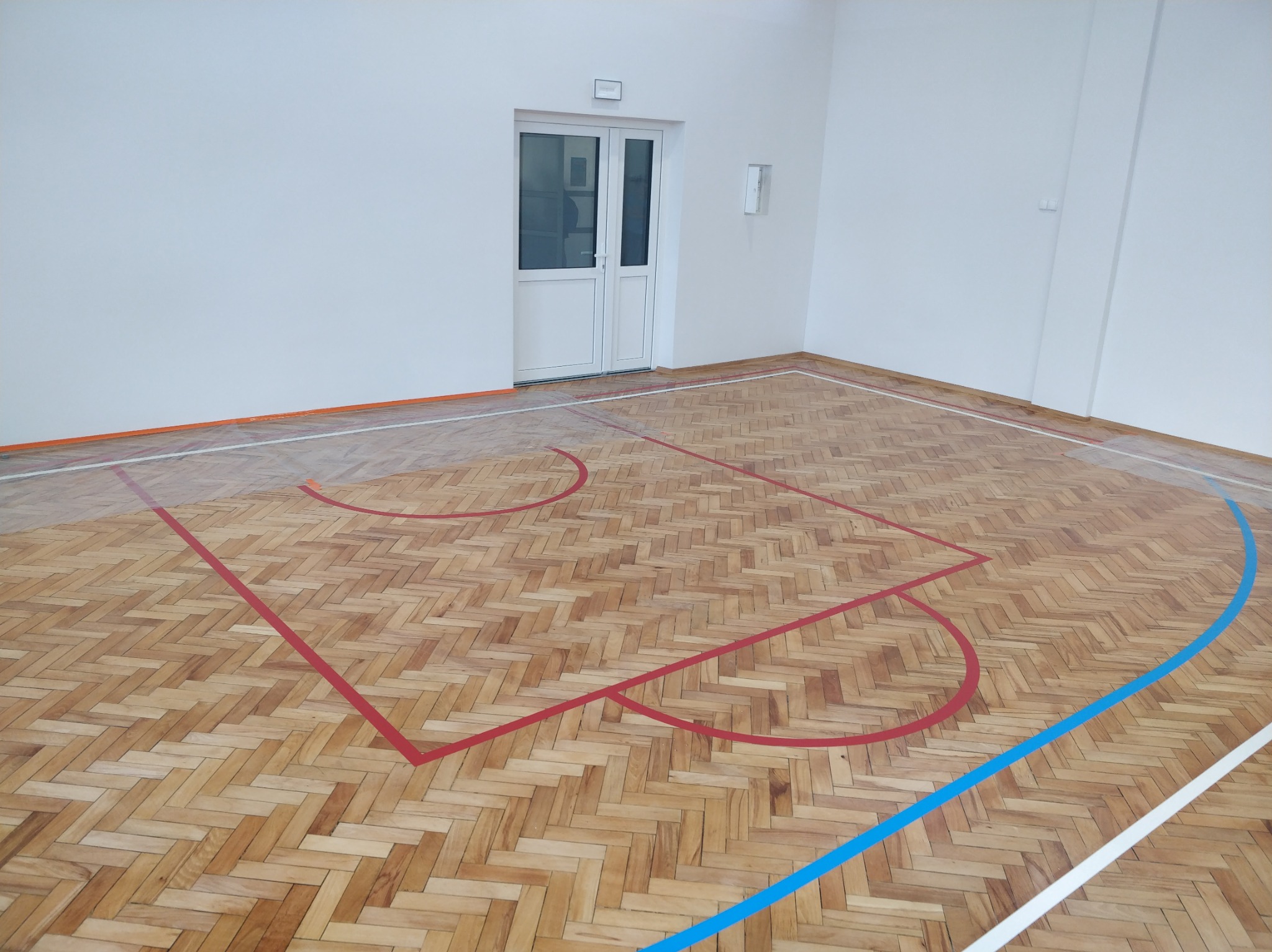 Parkiet w jodełkę na sali gimnastycznej z liniami boiska w kolorach czerwonym, niebieskim i białym. Widoczne drzwi i fragment ściany.