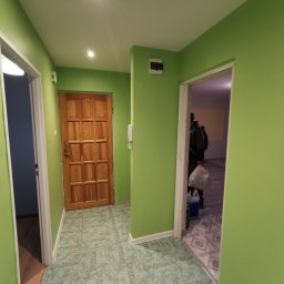 TP REMONTY I WYKOŃCZENIA - Korytarz w mieszkaniu z zielonymi ścianami, drewnianymi drzwiami i dwiema otwartymi przestrzeniami, w jednej z nich widoczna osoba z torbami.