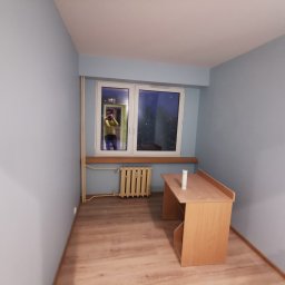 TP REMONTY I WYKOŃCZENIA - Pomalowane na jasnoniebiesko puste pomieszczenie z drewnianym biurkiem i grzejnikiem pod oknem, widoczny fotograf w odbiciu okna.