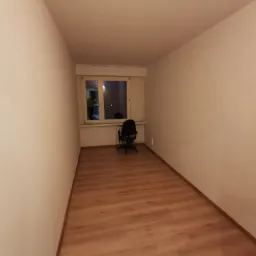 Pusty pokój z panelami podłogowymi, pomalowanymi na biało ścianami i oknem z widocznym krzesłem biurowym.