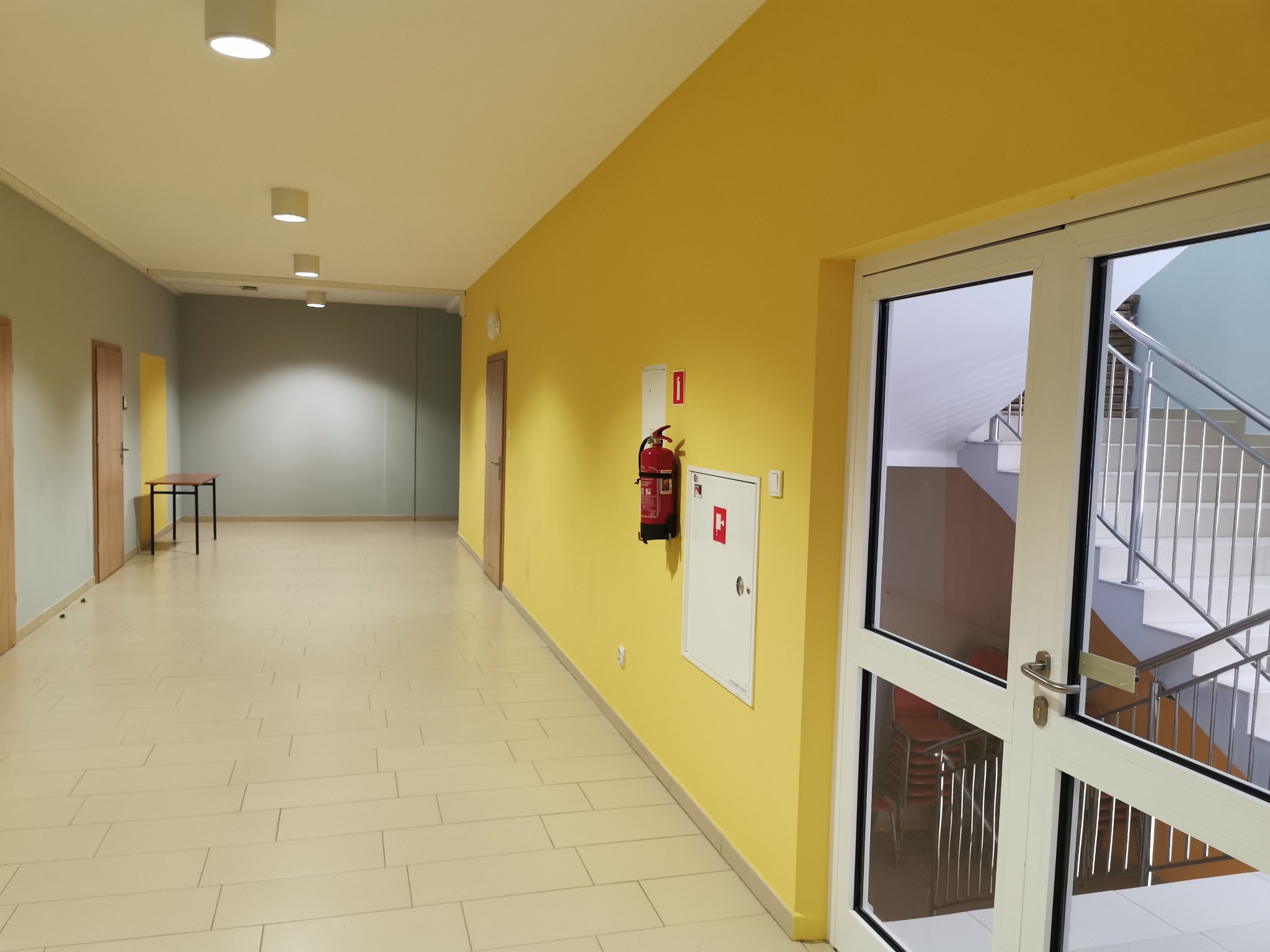 Korytarz w Sanoku z żółtą ścianą, białymi drzwiami z widokiem na klatkę schodową oraz widoczną gaśnicą na ścianie.