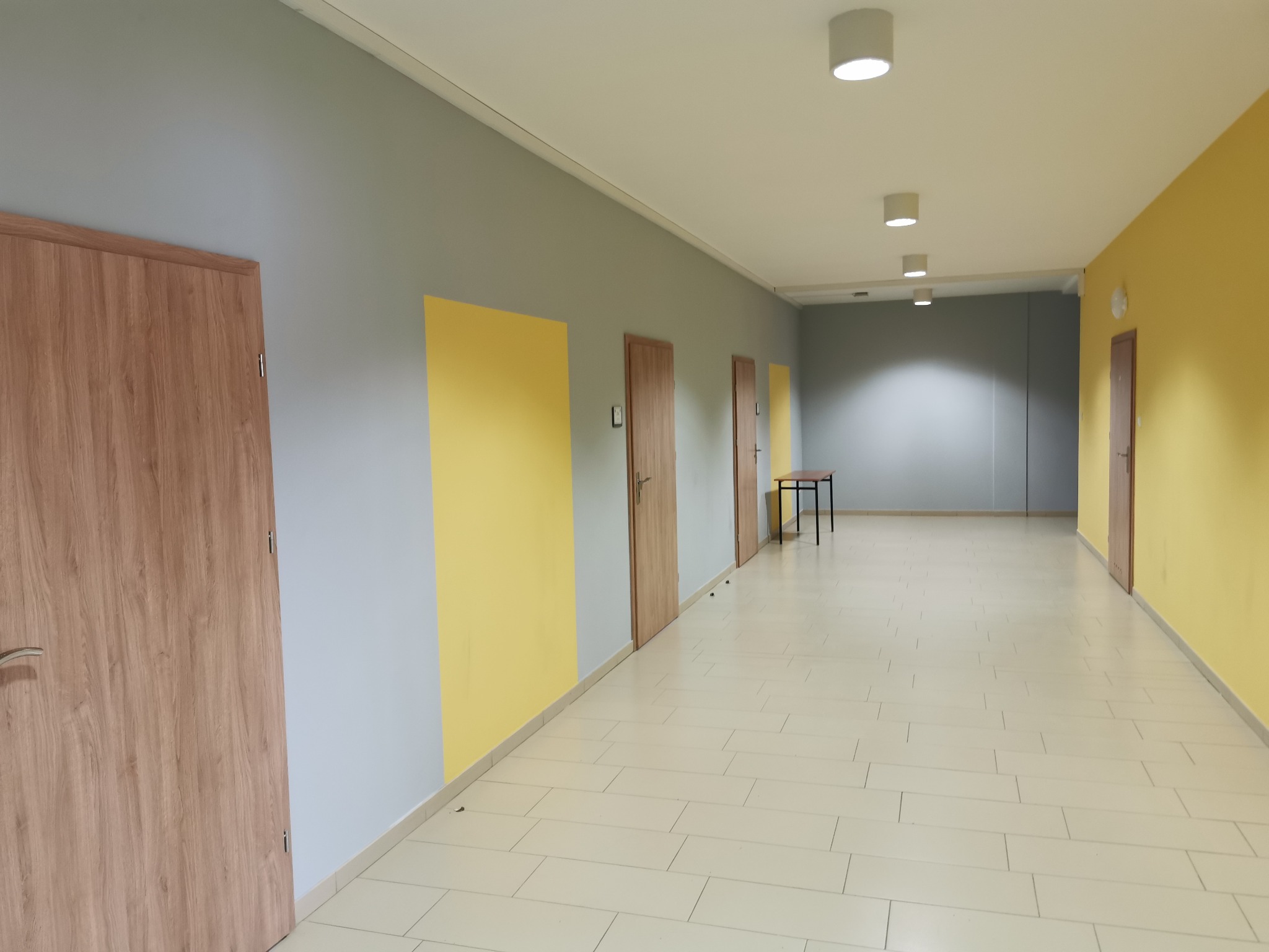 Długi korytarz z jasną podłogą, żółtymi i szarymi ścianami, oraz drewnianymi drzwiami po obu stronach, oświetlony rzędem okrągłych lamp sufitowych.