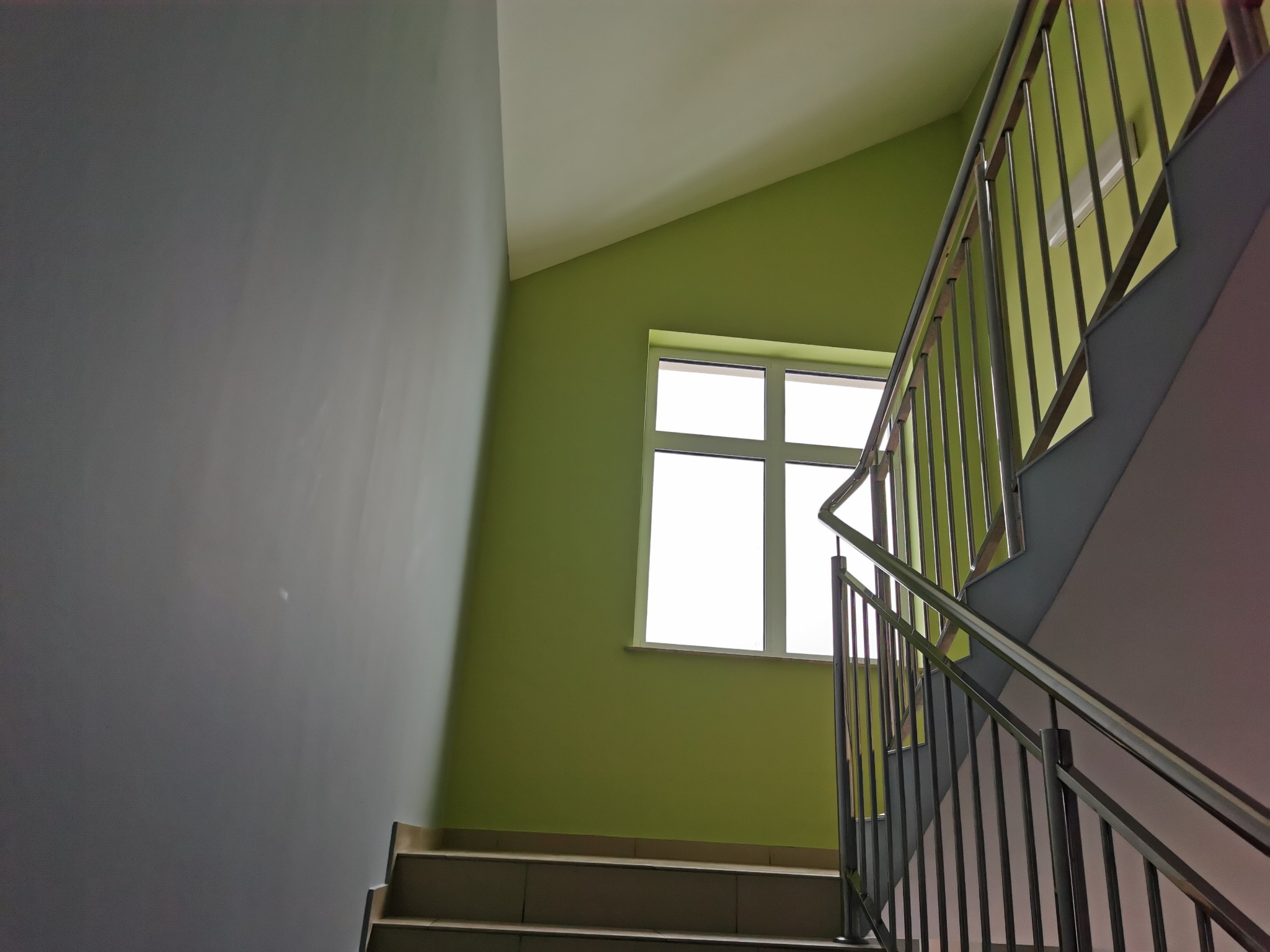 Klatka schodowa z metalową balustradą, ściany pomalowane na zielono i szaro, okno z białymi ramami w tle.