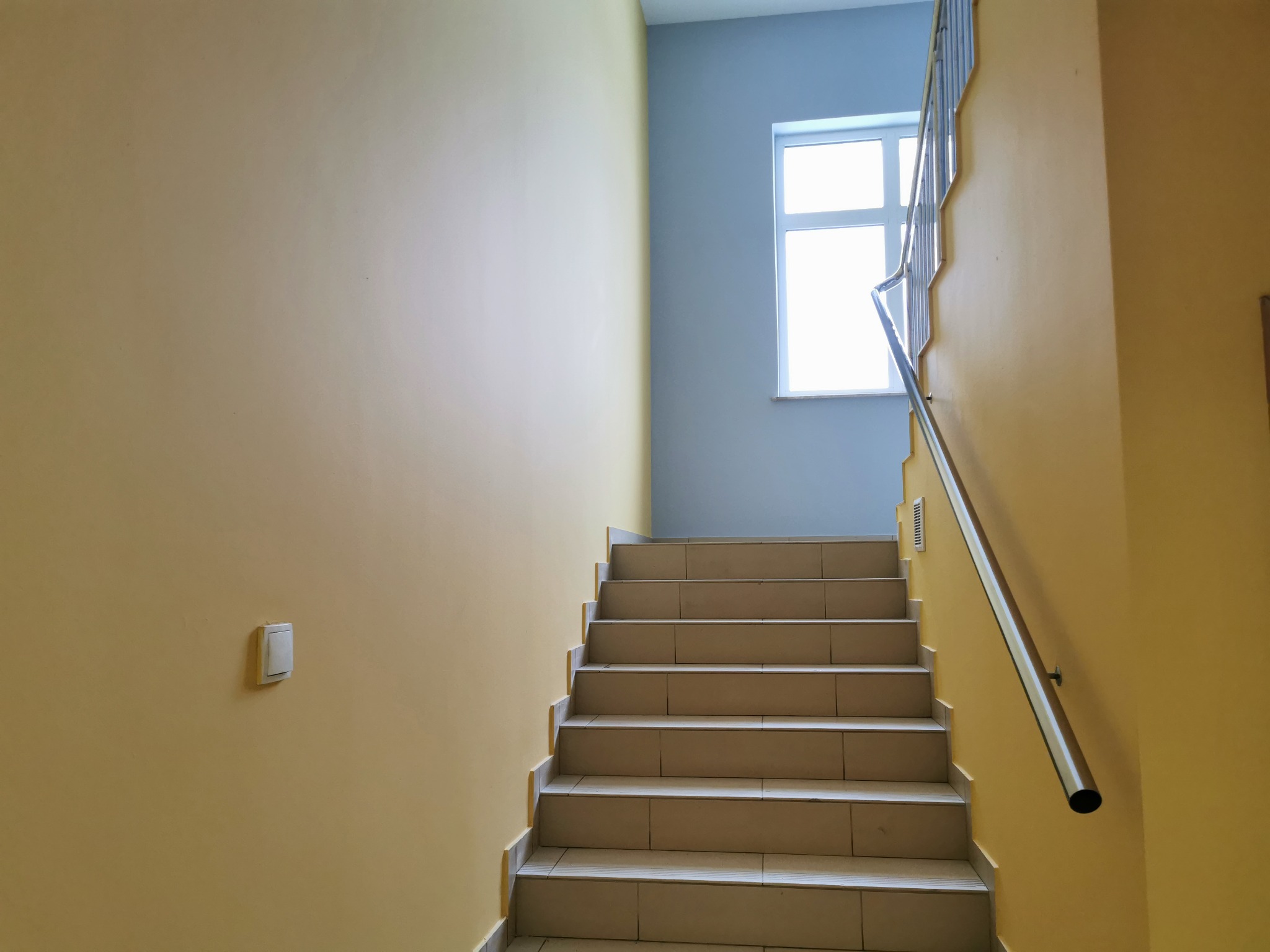 Klatka schodowa z jasnymi, kafelkowymi schodami, żółtymi ścianami i srebrną poręczą, oświetlona naturalnym światłem z okna na półpiętrze.
