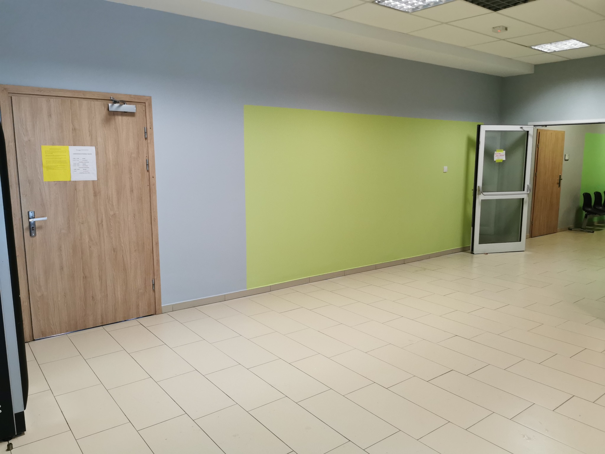 Pomalowane ściany w biurze na szaro i jasnozielono, z dwiema drewnianymi futrynami drzwi i białą futryną, kremowa podłoga z płytek.