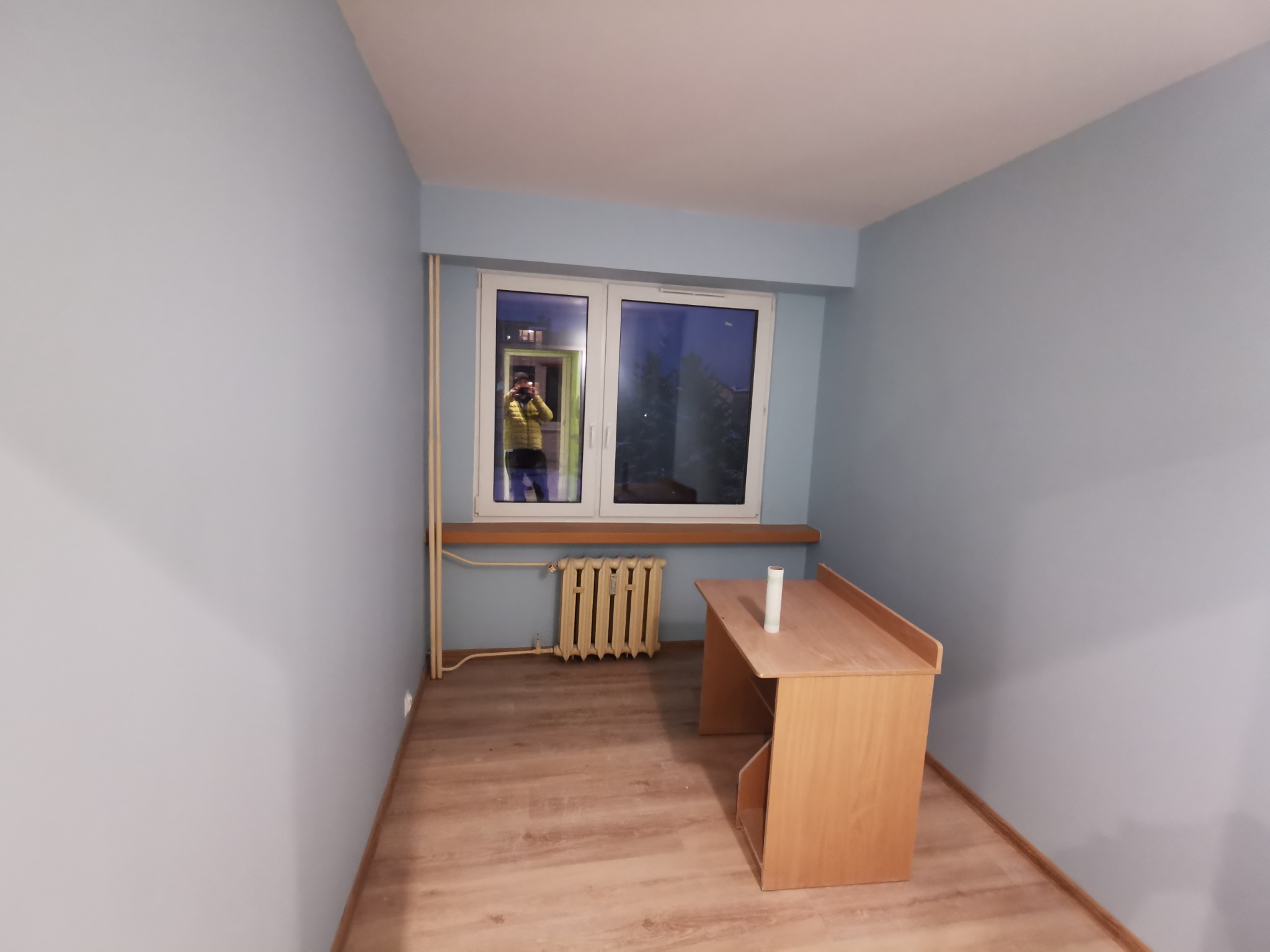 Pomalowane na jasnoniebiesko puste pomieszczenie z drewnianym biurkiem i grzejnikiem pod oknem, widoczny fotograf w odbiciu okna.