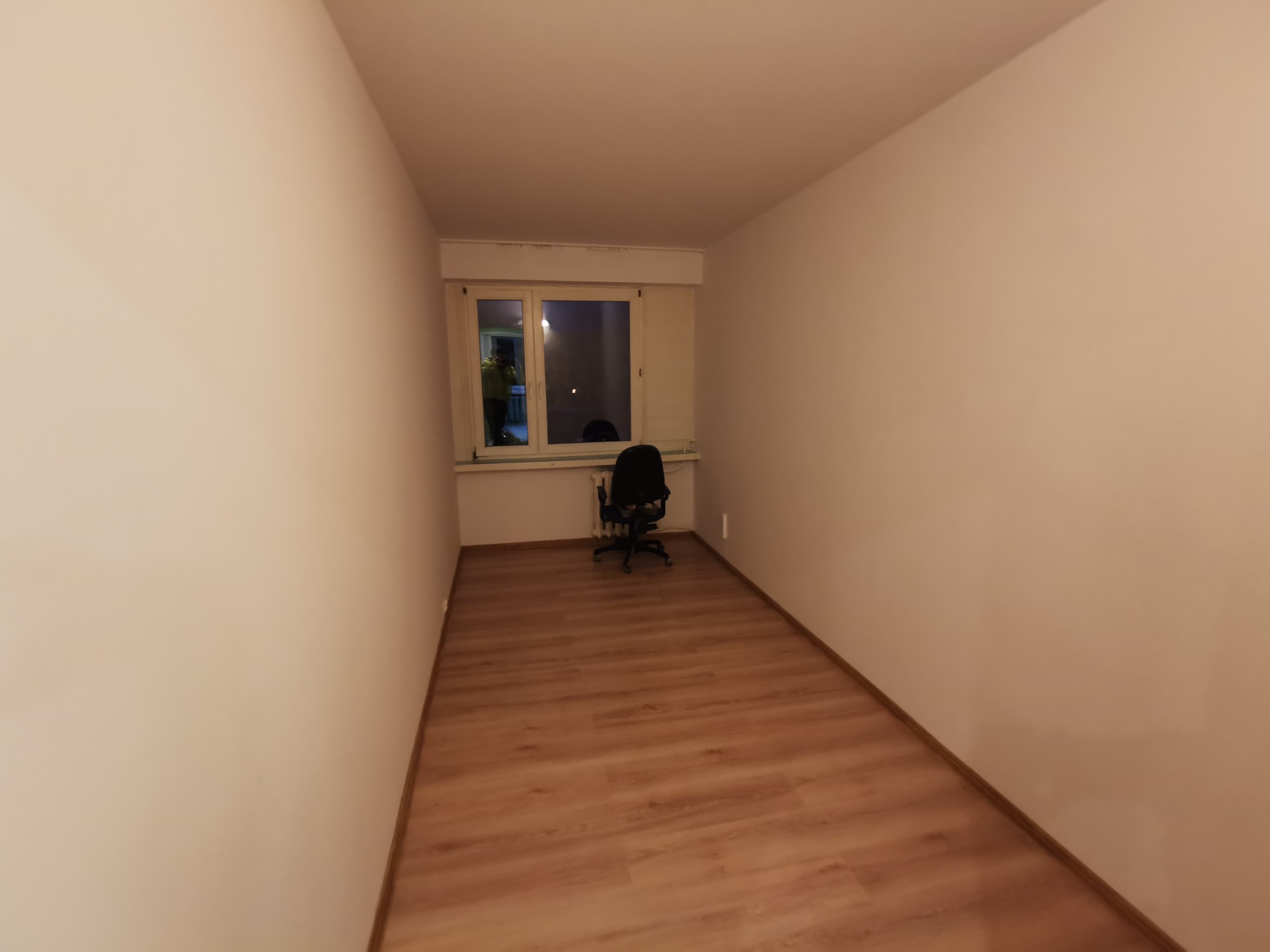 Pusty pokój z panelami podłogowymi, pomalowanymi na biało ścianami i oknem z widocznym krzesłem biurowym.